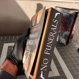 K&ouml;nnte beinhalten: Ein schwarzes Buch mit dem Titel "No Funerals" in wei&szlig; auf dem R&uuml;cken. Das Buch ist ge&ouml;ffnet und zeigt eine Seite aus dem Buch "Crooked Kingdom" von Leigh Bardugo.