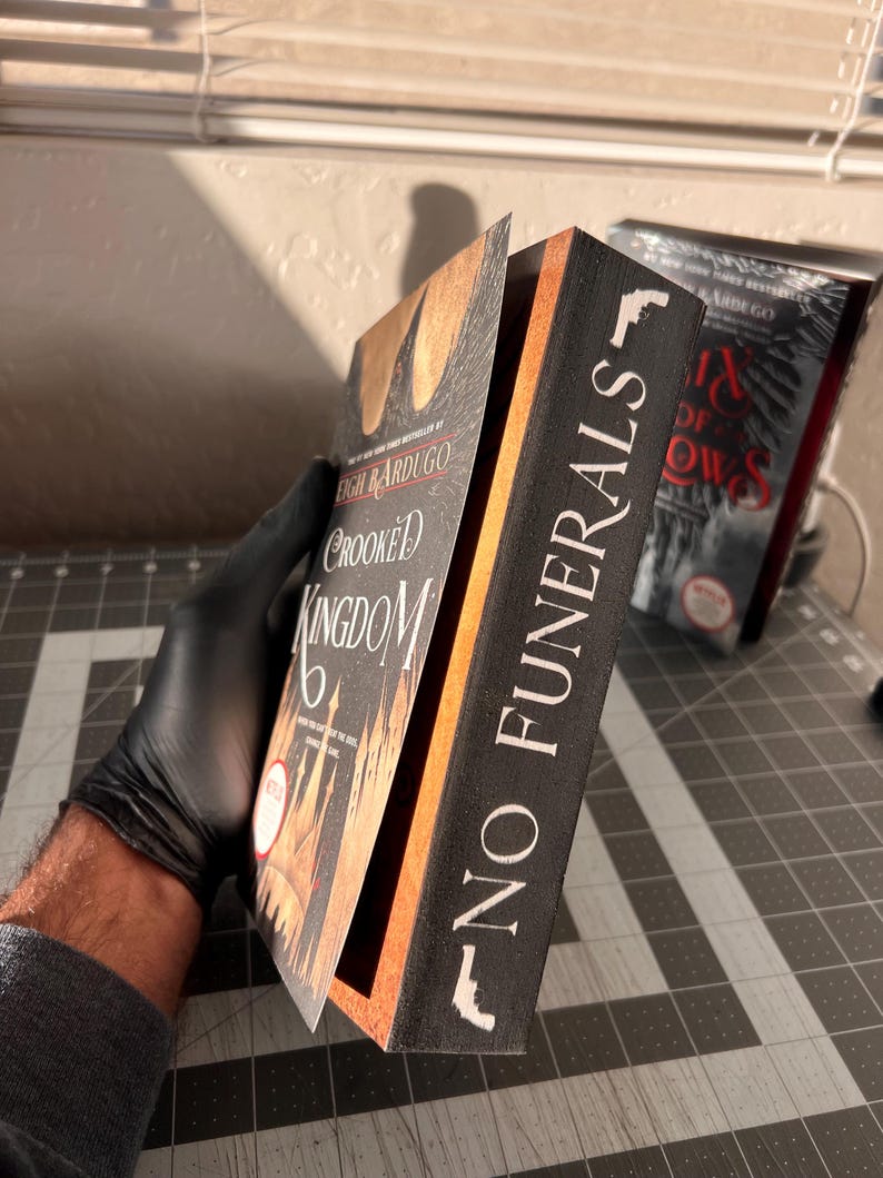 K&ouml;nnte beinhalten: Ein schwarzes Buch mit dem Titel "No Funerals" in wei&szlig; auf dem R&uuml;cken. Das Buch ist ge&ouml;ffnet und zeigt eine Seite aus dem Buch "Crooked Kingdom" von Leigh Bardugo.