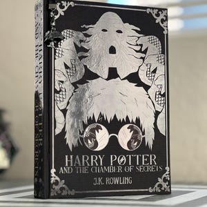 May include: A black book with a silver cover featuring a silver illustration of Harry Potter with a snake in his glasses. The title "Harry Potter and the Chamber of Secrets" is printed in silver on the cover, along with the author's name, J.K. Rowling.
