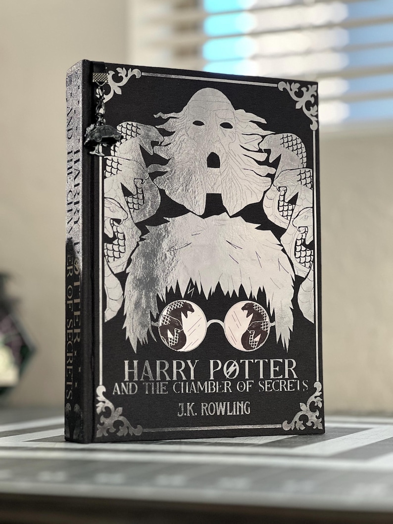 May include: A black book with a silver cover featuring a silver illustration of Harry Potter with a snake in his glasses. The title "Harry Potter and the Chamber of Secrets" is printed in silver on the cover, along with the author's name, J.K. Rowling.