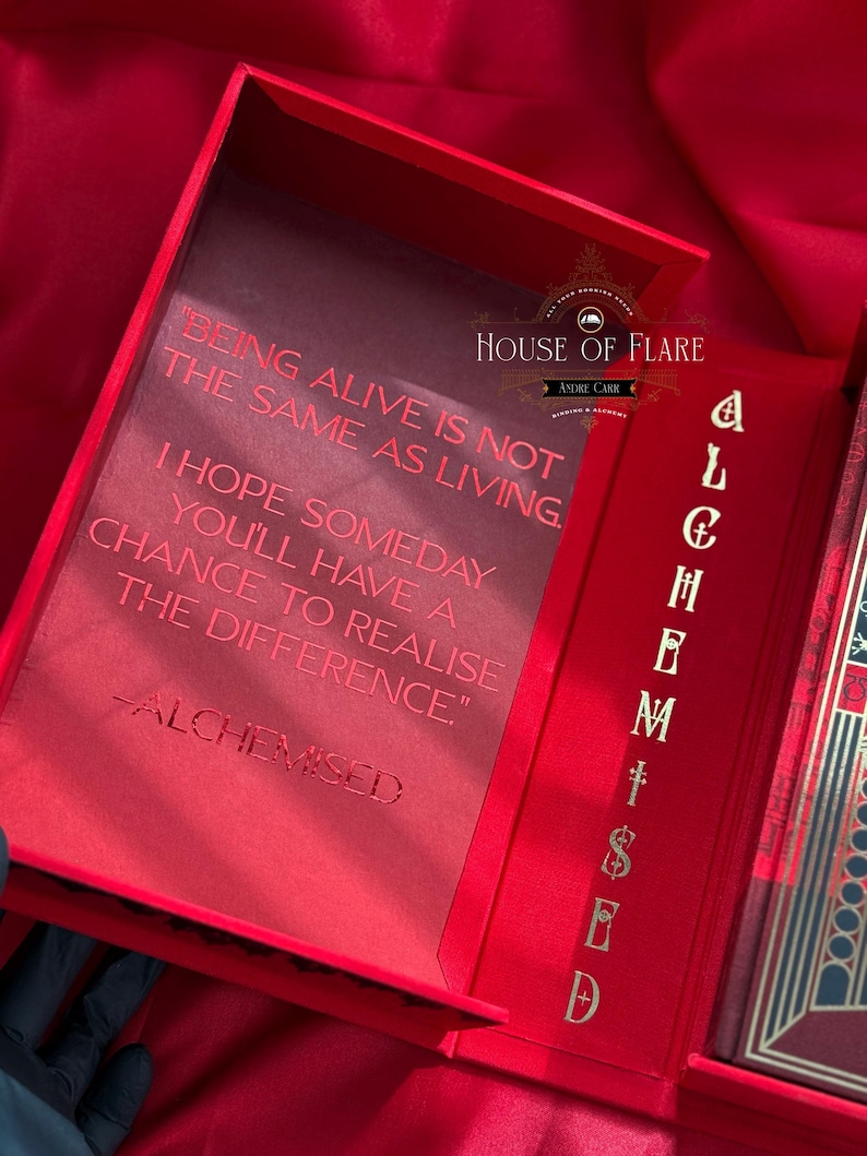 K&ouml;nnte beinhalten: Ge&ouml;ffnete rote Box mit goldfarbenen Buchstaben und dem Text "BEING ALIVE IS NOT THE SAME AS LIVING." Die Box hat auch den Text "ALCHEMISED" und "HOUSE OF FLARE". Die Box ist ge&ouml;ffnet und zeigt einen roten Innenraum.