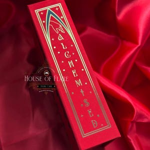 K&ouml;nnte beinhalten: Eine rote rechteckige Schachtel mit goldfarbenen Buchstaben, die "ALCHEMISED" lauten. Die Schachtel hat einen goldenen Rand und ein Design einer Hand und eines Torbogens. Die Schachtel befindet sich auf einem roten Satinhintergrund. Das Logo von House of Flare ist sichtbar.