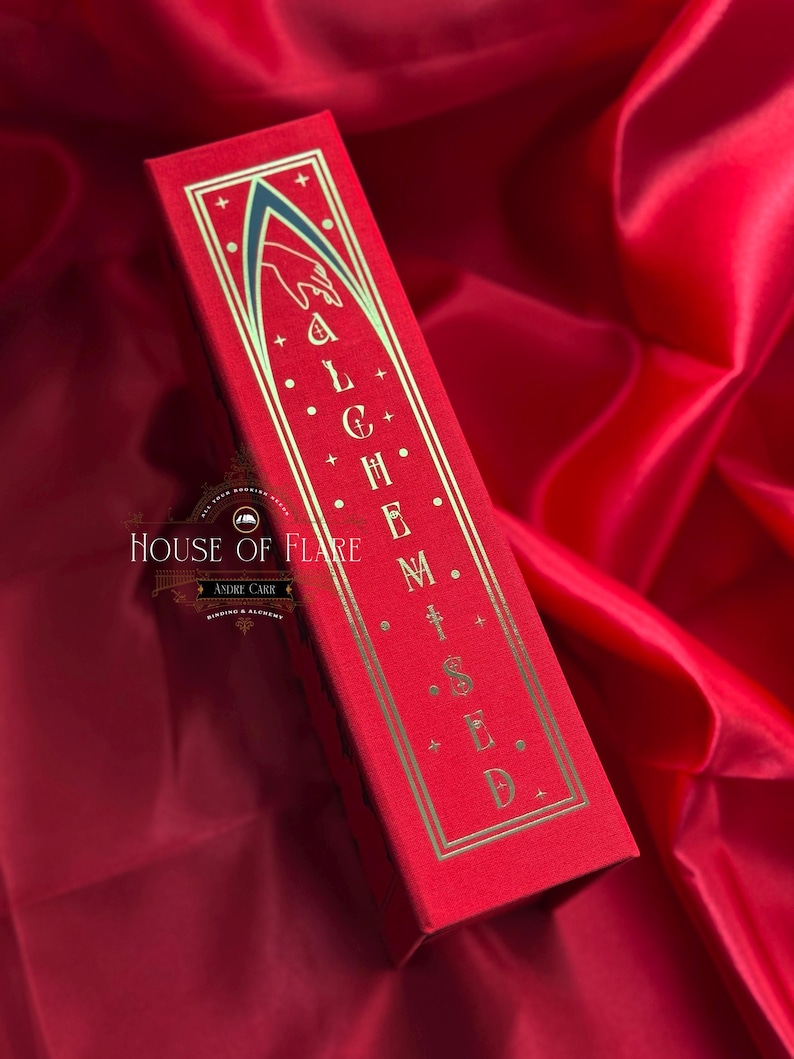 K&ouml;nnte beinhalten: Eine rote rechteckige Schachtel mit goldfarbenen Buchstaben, die "ALCHEMISED" lauten. Die Schachtel hat einen goldenen Rand und ein Design einer Hand und eines Torbogens. Die Schachtel befindet sich auf einem roten Satinhintergrund. Das Logo von House of Flare ist sichtbar.