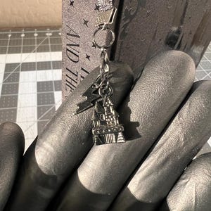 Puede incluir: Un charm plateado con un peque&ntilde;o castillo con un techo puntiagudo y una cadena unida a un anillo plateado. El charm cuelga de una bolsa de libro de tela gris con el texto "AND THE" y "ROWLI" visible.