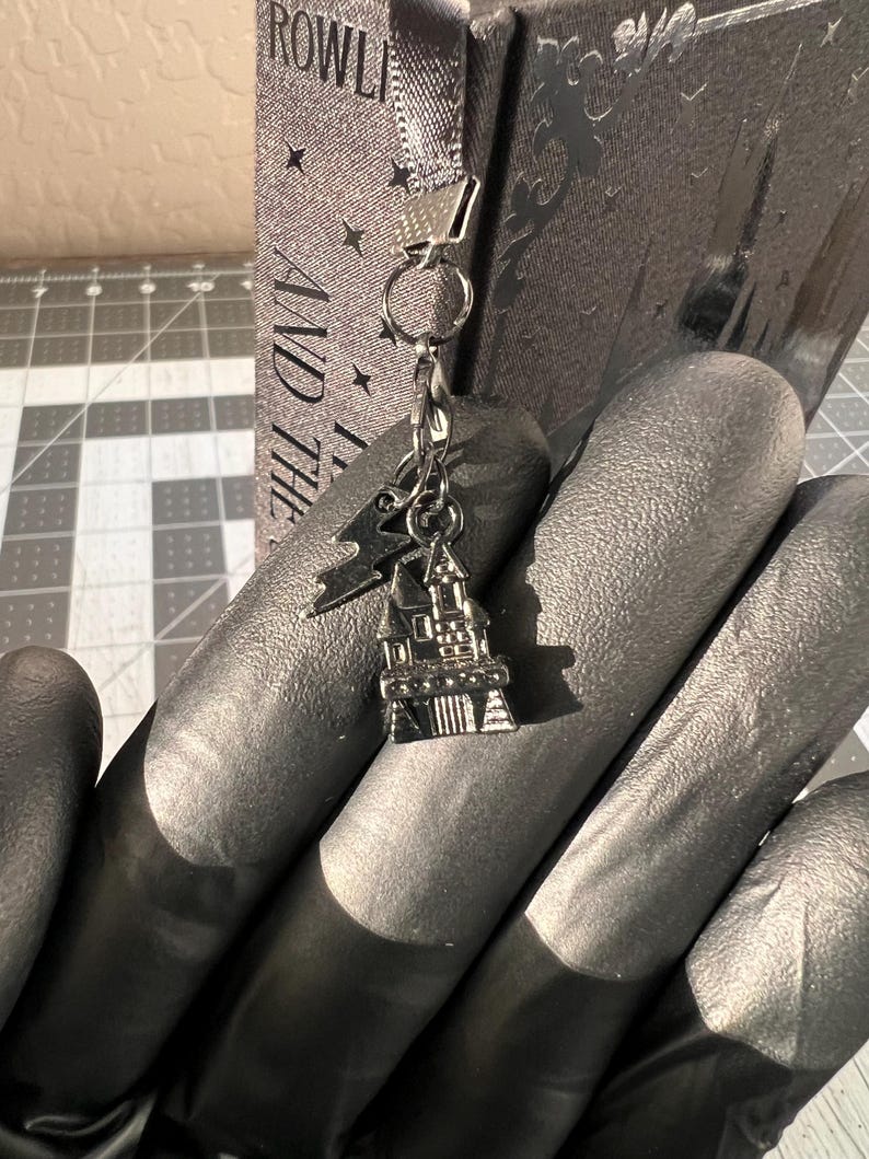 Puede incluir: Un charm plateado con un peque&ntilde;o castillo con un techo puntiagudo y una cadena unida a un anillo plateado. El charm cuelga de una bolsa de libro de tela gris con el texto "AND THE" y "ROWLI" visible.