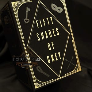 Może przedstawiać: Czarna książka ze złotymi akcentami i tytułem "Fifty Shades of Grey" złotymi literami. Okładka zawiera klucz, maskę i inne elementy dekoracyjne. Książka pochodzi z House of Flare.