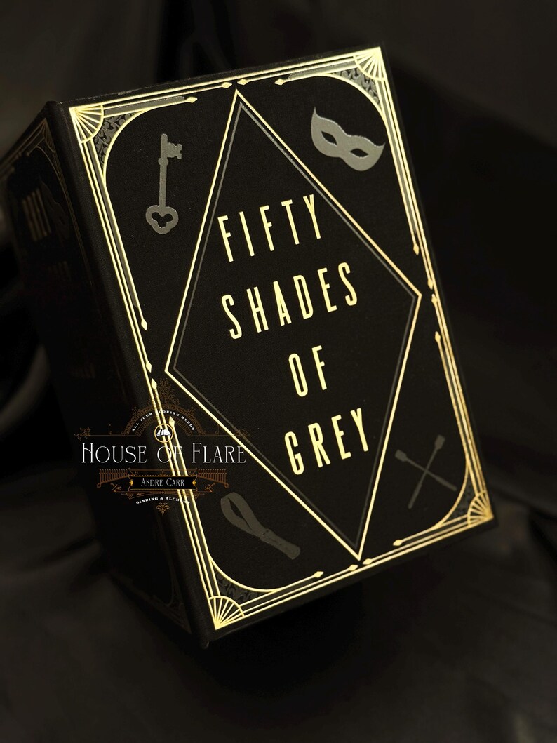 Może przedstawiać: Czarna książka ze złotymi akcentami i tytułem "Fifty Shades of Grey" złotymi literami. Okładka zawiera klucz, maskę i inne elementy dekoracyjne. Książka pochodzi z House of Flare.