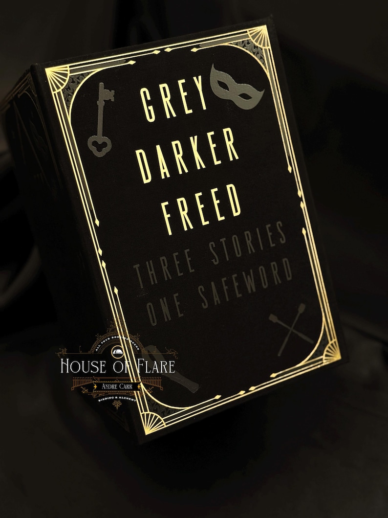 Może przedstawiać: Czarne pudełko ze złotym wykończeniem w stylu art deco i tekstem "GREY DARKER FREED THREE STORIES ONE SAFEWORD". Pudełko zawiera r&oacute;wnież klucz i maskę.