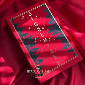 K&ouml;nnte beinhalten: Ein rotes Buch mit goldener Schrift und einem dekorativen Design. Das Cover zeigt das Wort "ALCHEMY" und eine Stadtsilhouette. Das Buch liegt vor einem roten Satinhintergrund, mit dem Text "HOUSE OF FLARE" unten.