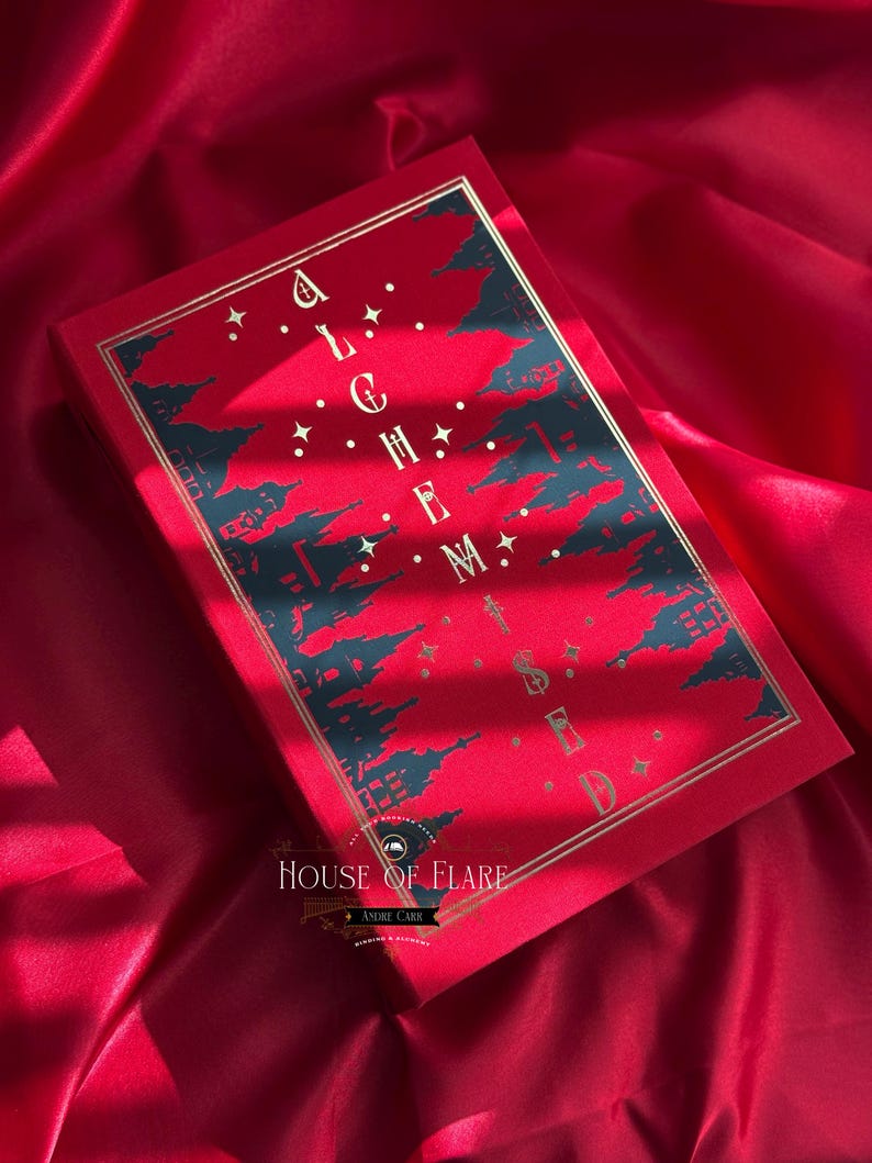 K&ouml;nnte beinhalten: Ein rotes Buch mit goldener Schrift und einem dekorativen Design. Das Cover zeigt das Wort "ALCHEMY" und eine Stadtsilhouette. Das Buch liegt vor einem roten Satinhintergrund, mit dem Text "HOUSE OF FLARE" unten.