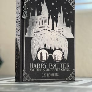 May include: A black hardcover book with a silver foil design of Hogwarts castle and Harry Potter. The title is "Harry Potter and the Sorcerer's Stone" by J.K. Rowling.