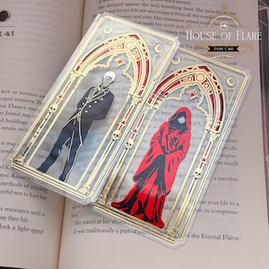 May include: Two rectangular clear acrylic bookmarks featuring gold foil gothic arch frames with skull accents. One bookmark displays a figure in a black suit, the other a figure in a red hooded cloak. Both include the text HOUSE OF FLARE ANDRE CARR.