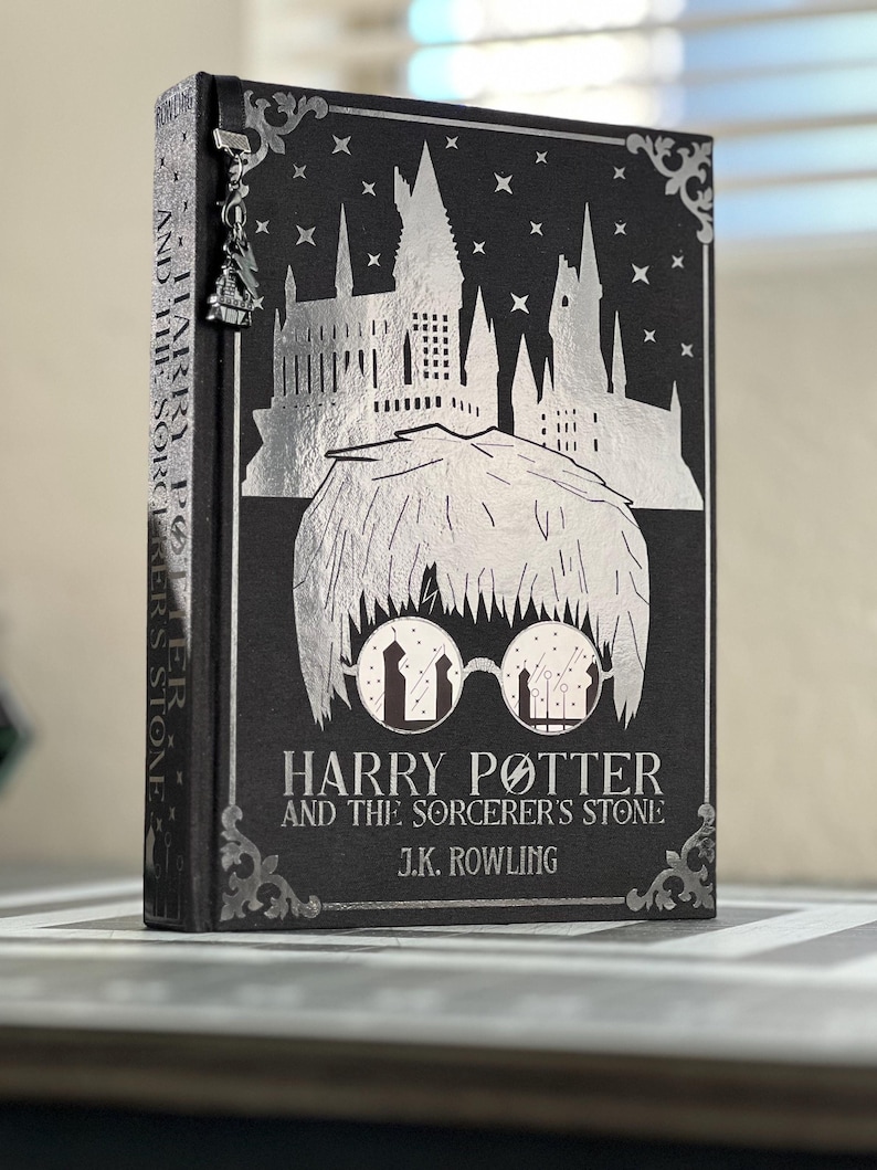 Puede incluir: Un libro negro de tapa dura con un dise&ntilde;o en plata de la escuela de magia de Hogwarts y Harry Potter con gafas. El t&iacute;tulo es "Harry Potter y la piedra filosofal" de J.K. Rowling.