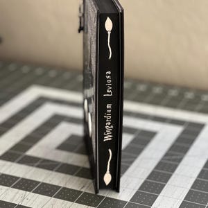 Puede incluir: Un libro negro con una ilustraci&oacute;n blanca de una varita m&aacute;gica y el texto "Wingardium Leviosa" en el lomo.