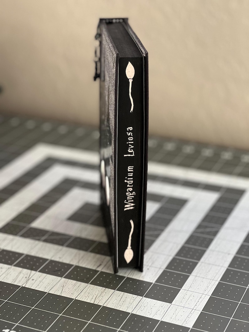 Puede incluir: Un libro negro con una ilustraci&oacute;n blanca de una varita m&aacute;gica y el texto "Wingardium Leviosa" en el lomo.