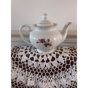 May include: A white porcelain teapot featuring a delicate pink rose floral pattern on the front. The teapot has a ribbed body, a curved handle, a matching lid with a finial, and gold trim accents along the rim, spout, and lid edge.
