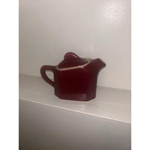 May include: A small, deep red ceramic creamer or teapot with a glossy finish, featuring a hexagonal body, a curved handle on the left side, a short spout on the right, and a matching lid with a small, pointed handle on top.