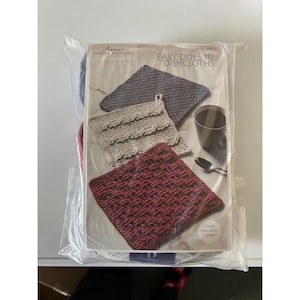 May include: A clear plastic package containing a crochet pattern book and yarn. The book cover reads "Annie's Caring Crochet Club" and "Easy Does It! Dishcloths." The dishcloths are shown in gray, cream, and burgundy. A cup and spoon are also visible.
