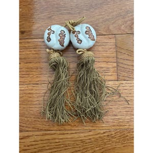 May include: Two decorative tassels featuring round, off-white ceramic beads with brown floral patterns. Each bead is topped with a gold-colored cord loop and a long, dense fringe of metallic gold-toned threads hanging from a matching gold-colored cap.