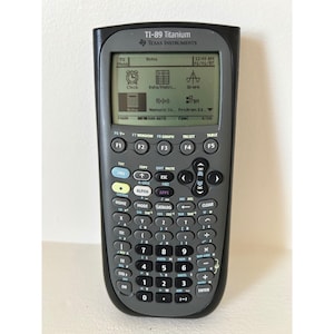 May include: A TI-89 Titanium calculator by Texas Instruments. The calculator has a black and gray design with a screen displaying various functions and options. It features a full keyboard with numerous buttons for mathematical operations and functions.