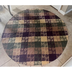 May include: A round tablecloth featuring a checkered pattern of alternating squares in shades of dark purple, olive green, and tan. Each square is decorated with a repeating leaf motif, creating a textured appearance across the entire fabric surface.