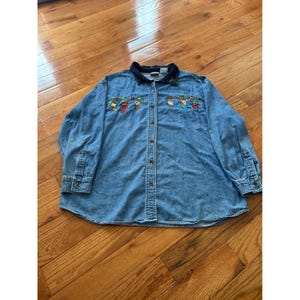 May include: A long-sleeve denim button-down shirt featuring a dark blue collar and embroidered holiday-themed designs on the chest. The embroidery depicts colorful stockings and greenery garlands on both sides of the front button placket.