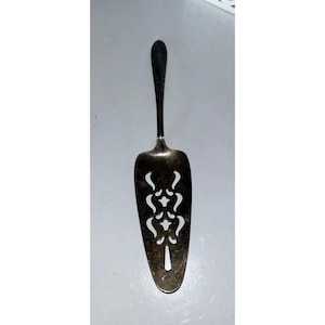 May include: A silver-toned pie server with a long handle and a slotted blade. The blade features a decorative cut-out design. The server is designed for serving pies, cakes, and other baked goods.