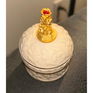 May include: A white, round decorative jar with a gold-colored lid and a red jewel. The jar has a leaf pattern and a gold rim. The lid features a detailed gold sculpture. The jar is likely made of porcelain or ceramic.