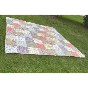 May include: A rectangular patchwork quilt featuring a grid of square fabric panels in various floral and botanical patterns. The color palette includes shades of sage green, dusty rose, lavender, and cream, arranged in a repeating geometric layout.