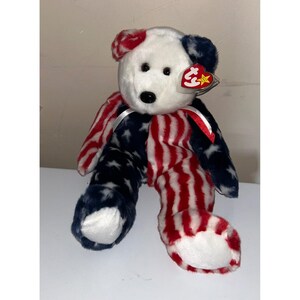 Peut inclure: Un ours en peluche patriotique avec une tête blanche et un corps rouge, blanc et bleu. Le corps de l'ours est divisé en sections à rayures rouges et blanches, et en sections bleues avec des étoiles blanches. Une étiquette rouge est attachée à l'oreille.