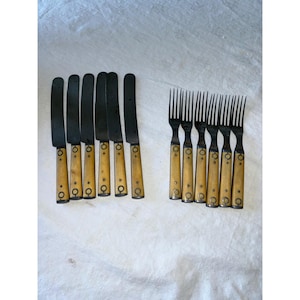May include: A set of antique cutlery, including eight knives and eight forks. The knives have black blades and light-colored handles with decorative metal accents. The forks have black tines and matching handles. The set is arranged on a white surface.