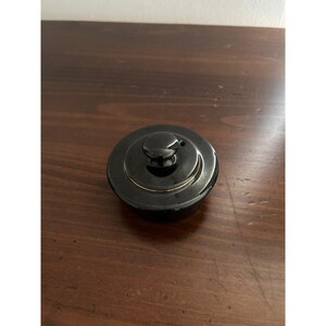 May include: A circular black ceramic lid featuring a central knob handle and a thin gold-colored rim accent. The lid has a smooth, glossy finish and a small circular vent hole positioned near the central knob on the top surface of the object.