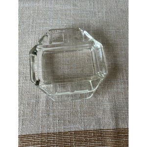 Puede incluir: Un cenicero de vidrio transparente, de forma octogonal. El cenicero tiene un interior rectangular con un borde ligeramente elevado y un pequeño labio en un lado. El vidrio es transparente, lo que permite ver la superficie que hay debajo. El cenicero es probablemente vintage.