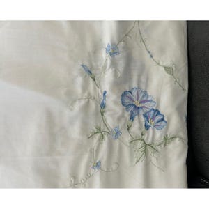 May include: A white fabric textile featuring delicate embroidery of blue morning glory flowers with green stems and leaves. The floral pattern is arranged in a climbing vine design that extends across the smooth, light-colored surface of the material.
