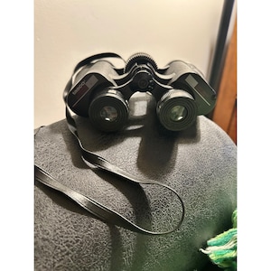 May include: Black Tasco binoculars with a black neck strap. The binoculars have a textured surface and are resting on a dark gray surface. The word "tasco" is printed on the side of the binoculars.