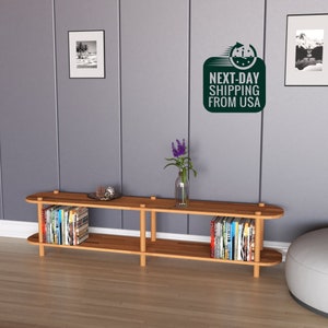 May include: A light wood shelf unit with two shelves and rounded edges. The shelf is filled with books and has a vase of purple flowers on top. The shelf is against a gray wall with a framed black and white photo and a sticker that says "NEXT-DAY SHIPPING FROM USA".