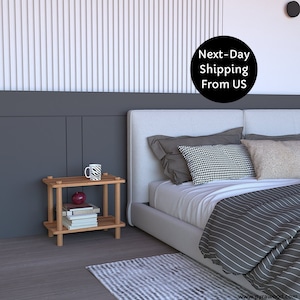 May include: A wooden two-tiered end table with a white mug and a red apple on the top shelf. Books are stacked on the bottom shelf. The table is next to a white bed with gray and white striped bedding.