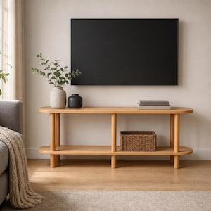 May include: A light wood console table with rounded ends and two tiers, featuring a beige textured vase with green foliage, a small black vase, two stacked gray books, and a woven brown rectangular basket on the lower shelf below a wall-mounted TV.