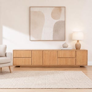 May include: A long, rectangular light wood media console with four drawers and two cabinet doors. On top sits a table lamp with a beige base and white shade, and a small, round, light-colored ceramic vase. Above hangs a square abstract wall art piece.