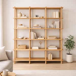 May include: A light-colored wooden bookshelf with multiple shelves. The shelves are filled with books, decorative vases, bowls, and a small camera. A potted plant is to the right of the bookshelf. The bookshelf is a light natural wood color.