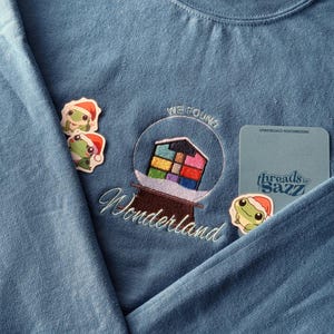 Può includere: Una felpa blu con testo ricamato che recita "We Found Wonderland" e un cubo di Rubik colorato all'interno di una cupola di vetro. Ci sono tre adesivi di rane sulla felpa, uno dei quali indossa un cappello di Babbo Natale.