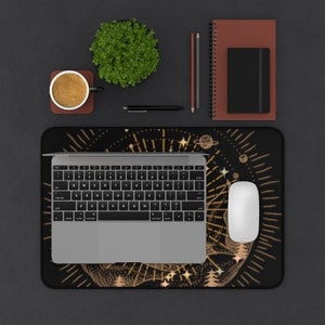 May include: A black mouse pad with a gold and white celestial design. The design features stars, planets, and a sun with rays. The mouse pad is on a black desk with a laptop, a coffee cup, a plant, and pens.