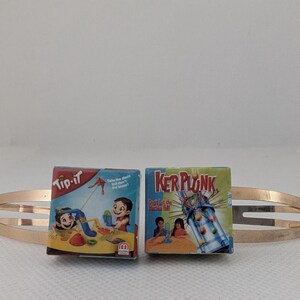 May include: A gold-toned metal hair clip holds two miniature game boxes. One box is labeled "Tip-It" with a beach scene, and the other is "Ker Plunk" with a colorful game illustration. The clip is a simple, elongated oval shape.