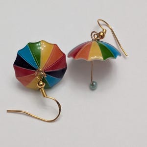 May include: A pair of gold dangle earrings with rainbow colored umbrellas. The umbrellas have a white center and are accented with a small blue bead.