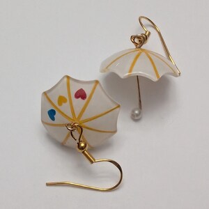 May include: A pair of gold earrings with white and yellow umbrella charms. The umbrellas have small painted hearts in blue, yellow, and red.