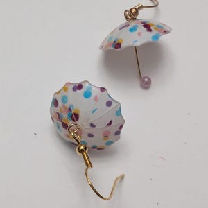 May include: A pair of gold earrings with a white umbrella charm. The umbrella has a colorful confetti pattern with blue, pink, purple, and yellow dots.