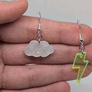 May include: A pair of earrings with a white cloud charm and a green lightning bolt charm. The earrings are silver and have a hook closure.