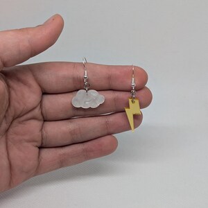 May include: A pair of mismatched earrings. One earring is a white cloud shape, and the other is a gold lightning bolt. Both earrings have silver-colored fish hook closures. The earrings are displayed against a neutral background.