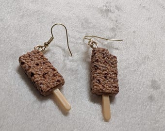 May include: A pair of novelty earrings shaped like chocolate-covered ice cream bars on a stick. The earrings have a textured brown surface with dark brown speckles, and a light beige stick. The earring hooks are gold-colored.