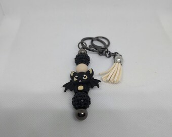 May include: A black bat charm with yellow eyes and wings, attached to a black and silver beaded keychain with a white tassel.